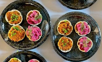 Black plates hold small tartlets with colourful fillings: some with a bright pink mixture topped with dill, others with an orange-brown mix garnished with green herbs, arranged neatly on a white surface.
