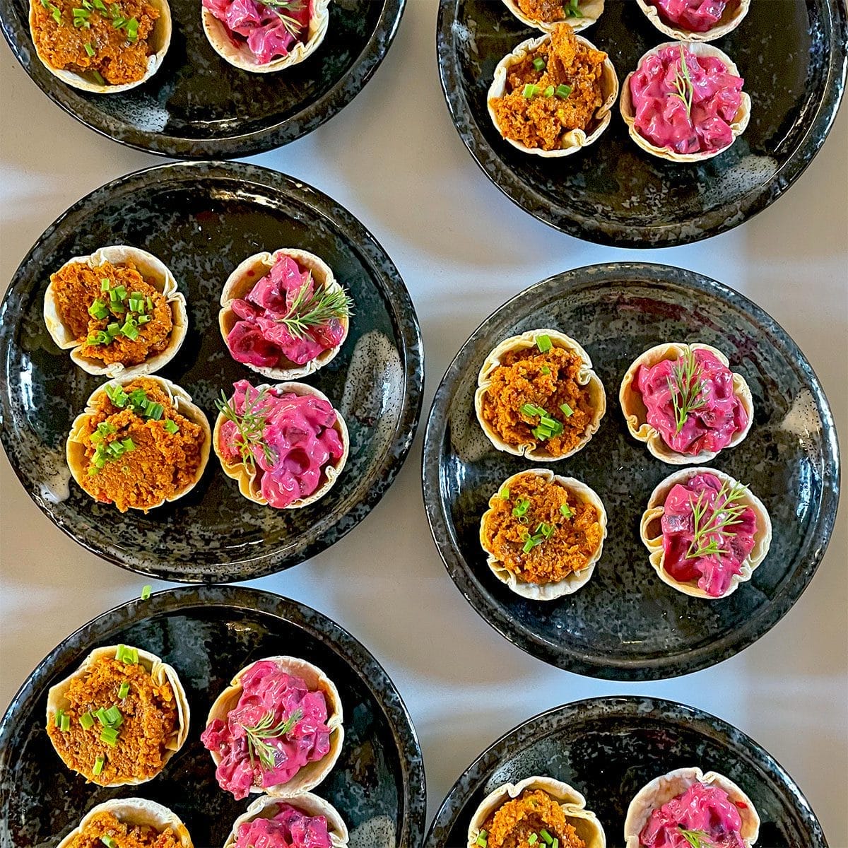 Black plates hold small tartlets with colourful fillings: some with a bright pink mixture topped with dill, others with an orange-brown mix garnished with green herbs, arranged neatly on a white surface.