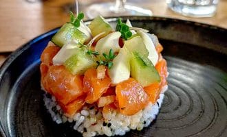 A round serving of sushi rice topped with diced salmon, avocado slices, diced cucumber, and microgreens, presented on a black plate with a glass of water in the background.