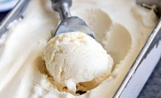 A metal ice cream scoop with a wooden handle is scooping vanilla ice cream from a rectangular metal container filled with smooth, creamy ice cream.
