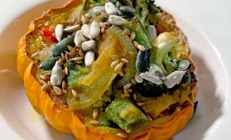 A baked orange squash half filled with roasted vegetables, topped with sunflower, pumpkin, and sesame seeds, served on a white plate.
