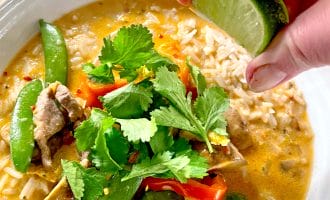 A hand squeezes a lime wedge over a bowl of rice and curry with vegetables, garnished with fresh coriander.