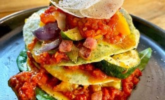 A stack of courgette slices layered with tomato sauce, diced vegetables, and pancetta, topped with grated cheese and black pepper, served on a dark plate with spinach leaves.
