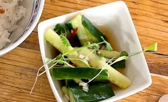 A small white bowl with cucumber slices, green herbs, and microgreens sits on a wooden table next to a bowl of white rice.