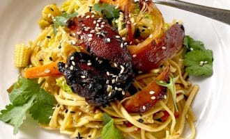 A white plate with stir-fried noodles, glazed vegetables, charred pieces of vegan mock duck, baby corn, carrots, fresh coriander leaves, and sesame seeds, with a fork resting on the edge.