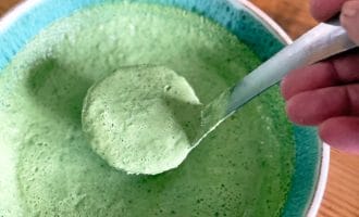 A hand holds a spoon scooping thick, green sauce or dressing from a turquoise-rimmed bowl on a wooden table. The sauce appears creamy and well-blended.