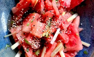 A bowl of watermelon pieces mixed with thinly sliced vegetables, topped with sesame seeds and chopped herbs, served in a blue ceramic dish.