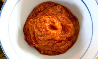 A white bowl with a blue rim filled with a thick, reddish-orange paste or sauce, placed on a wooden surface.