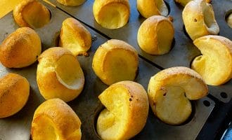 Golden-brown Yorkshire puddings rise and puff out of a black muffin tray, fresh from the oven, sitting on a wooden worktop.