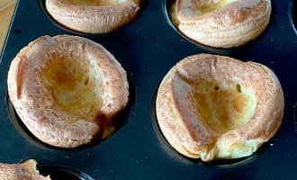 A close-up of freshly baked Yorkshire puddings in a muffin tray, showing their golden-brown, puffy edges and hollow centres.
