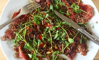 A plate of thinly sliced beef carpaccio topped with microgreens, sesame seeds, and chopped red peppers, with a fork and knife resting on the plate.