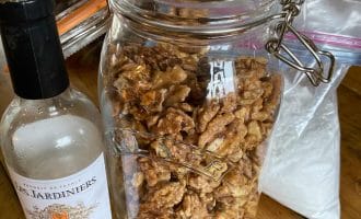 A large glass jar filled with candied walnuts sits on a wooden table next to a partially empty bottle of white wine. A plastic bag is in the background.