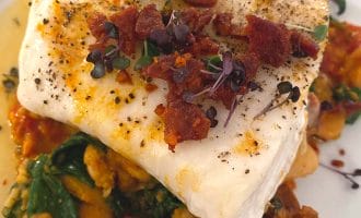 A fillet of white fish topped with microgreens and crispy bacon bits, served on a bed of wilted spinach, beans, and a tomato-based sauce on a white plate.