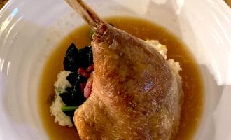 A cooked duck leg served on a bed of mashed potato and sautéed greens, surrounded by a pool of brown sauce on a white plate.