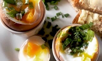 Two soft-boiled eggs in egg cups, topped with chopped spring onions and salt, with egg soldiers (buttered toast strips) and extra spring onions on a white plate. The yolks are runny and vibrant yellow.