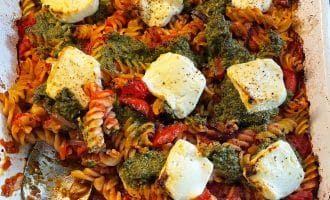 A baked pasta dish with spiral pasta, tomato sauce, dollops of green pesto, and several slices of melted white cheese on top, served in a white rectangular baking dish with a large serving spoon.