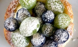 A pastry topped with green and purple grapes and dusted with icing sugar, placed on a white plate.