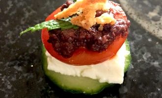 A cucumber slice topped with a layer of white cheese, a cherry tomato slice, olive tapenade, two mint leaves, and crispy fried onions, arranged on a dark, textured surface.