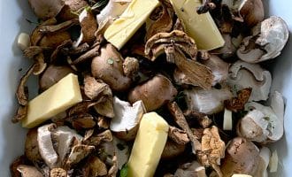 A close-up of raw mushrooms, both whole and sliced, mixed with dried mushrooms, sprigs of fresh herbs, and rectangular pieces of butter in a white dish.