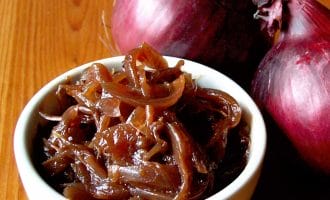 A small white bowl filled with caramelised onions sits on a wooden surface next to two whole red onions.