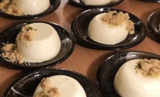 Several black plates, each with a serving of creamy white panna cotta topped with a small crumble and herbs, are arranged on a wooden surface, ready to be served.