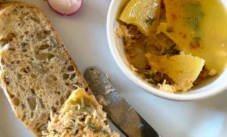 A slice of multigrain bread spread with a salmon pâté sits on a plate next to a bowl of pâté topped with jelly, a butter knife, and slices of fresh radish.