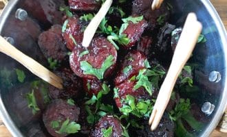 A metal bowl filled with glazed sausage slices garnished with chopped parsley, with several wooden sticks inserted for serving, placed on a wooden surface.