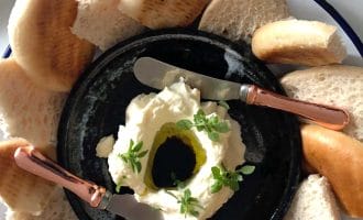 A black plate with a creamy white dip topped with olive oil and herbs, surrounded by slices of pitta bread, and two rose-gold handled spreading knives resting on the plate.