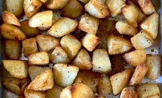 Golden brown roasted potato cubes spread out on a baking tray, seasoned with salt and pepper, showing crispy edges and a slightly oily surface.
