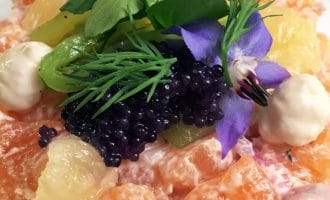 A colourful gourmet salad with orange segments, black caviar, citrus pieces, green herbs, edible flowers, and dollops of creamy sauce, artfully arranged on a white plate.
