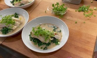 A bowl with fish fillet on a bed of greens, topped with fresh herbs and sauce. Two more similar bowls and a container of herbs are on a wooden worktop with chopped spring onions.