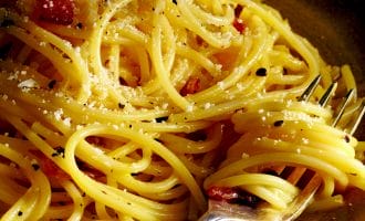 A close-up of cooked spaghetti on a plate, sprinkled with grated cheese and cracked black pepper, with a fork twirling some noodles.