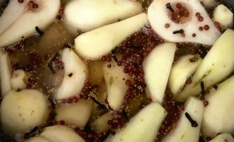 Sliced pears soaking in a saucepan with whole cloves, mustard seeds, and pink peppercorns, possibly being prepared for pickling or poaching.