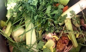 A pot filled with various fresh vegetables and herbs, including leeks, celery, carrots, parsley, thyme, and chives, arranged together and ready for cooking, possibly to make vegetable stock or broth.