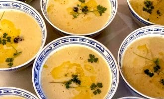 Several bowls of creamy yellow soup are garnished with fresh herbs and a drizzle of oil, served in blue and white patterned bowls, arranged closely together on a table.