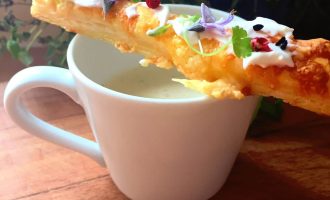 A white cup filled with creamy soup is topped with a long, flaky pastry stick garnished with cream, microgreens, peppercorns, and edible flowers, set on a wooden surface with green plants in the background.