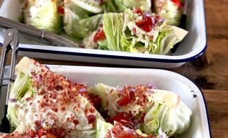 Trays of wedge salads topped with creamy dressing, diced tomatoes, bacon pieces, chopped red onion, and chives, served on white enamel trays with metal tongs on a wooden table.